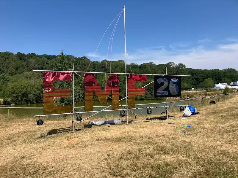 A large sign that reads EMF 26, made out of a shiny metallic material, mounted on scaffolding, next to a lake.